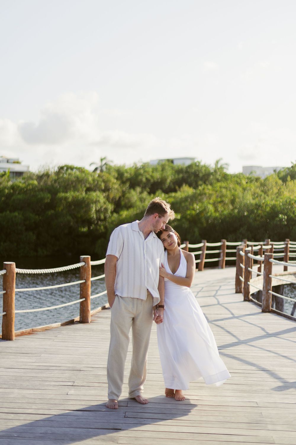 Couple in white outfits sharing a tender moment on a wooden boardwalk surrounded by lush tropical greenery.