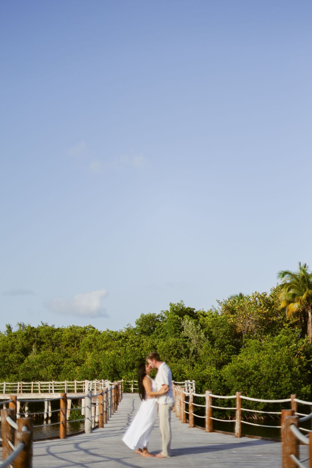 Couple kissing on a wooden boardwalk surrounded by tropical trees under a bright blue sky.