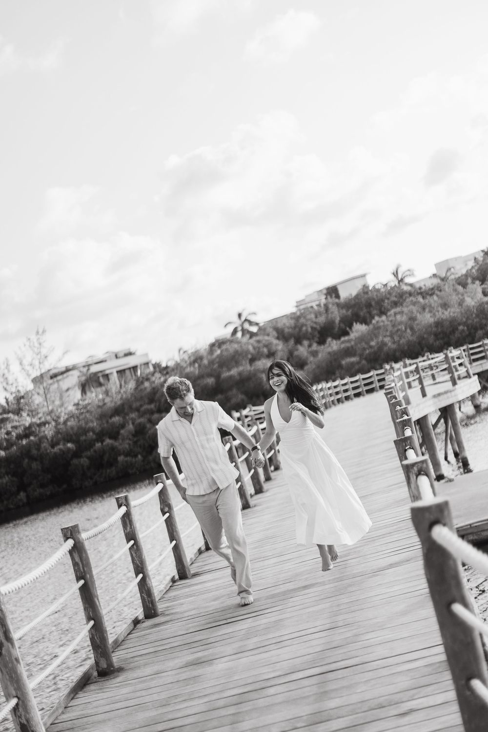 Black and white photo of a couple walking hand in hand on a wooden pier over water.