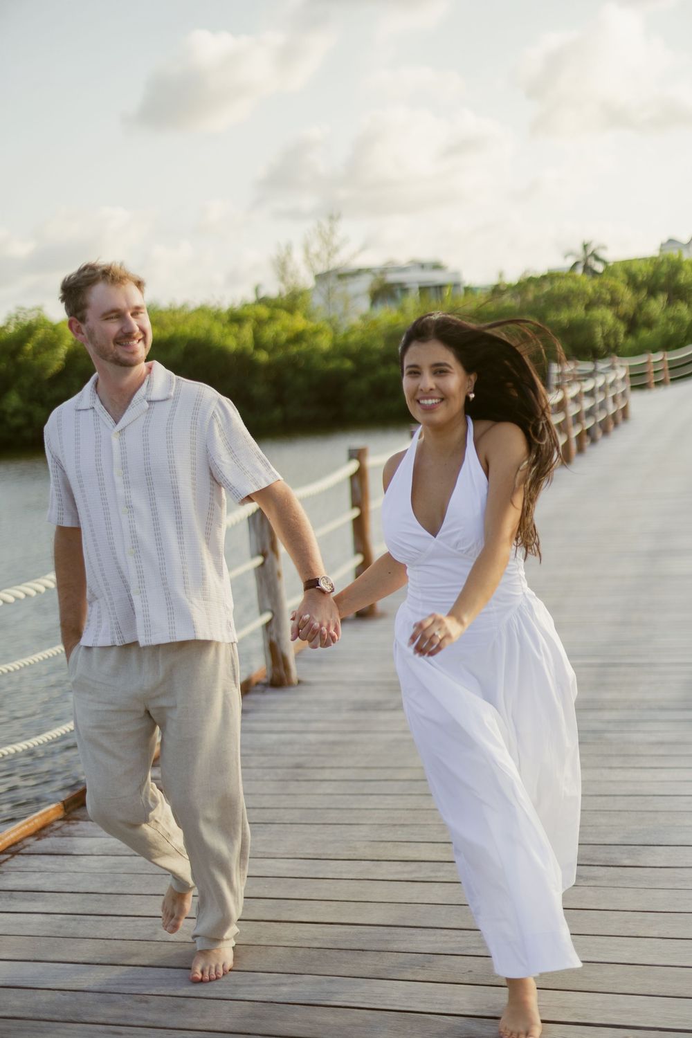 Smiling couple in white outfits holding hands while walking barefoot on a sunny tropical boardwalk.