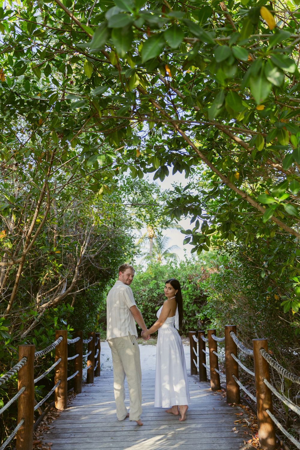 Couple holding hands on a wooden boardwalk framed by a lush tropical tree canopy.