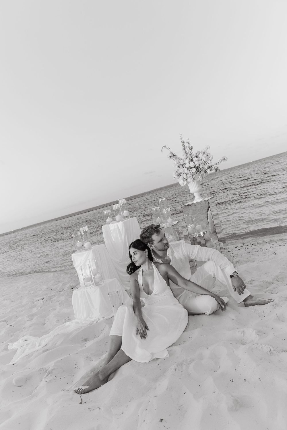Black and white photo of couple sitting on beach sand with floral arrangement and candles behind them.
