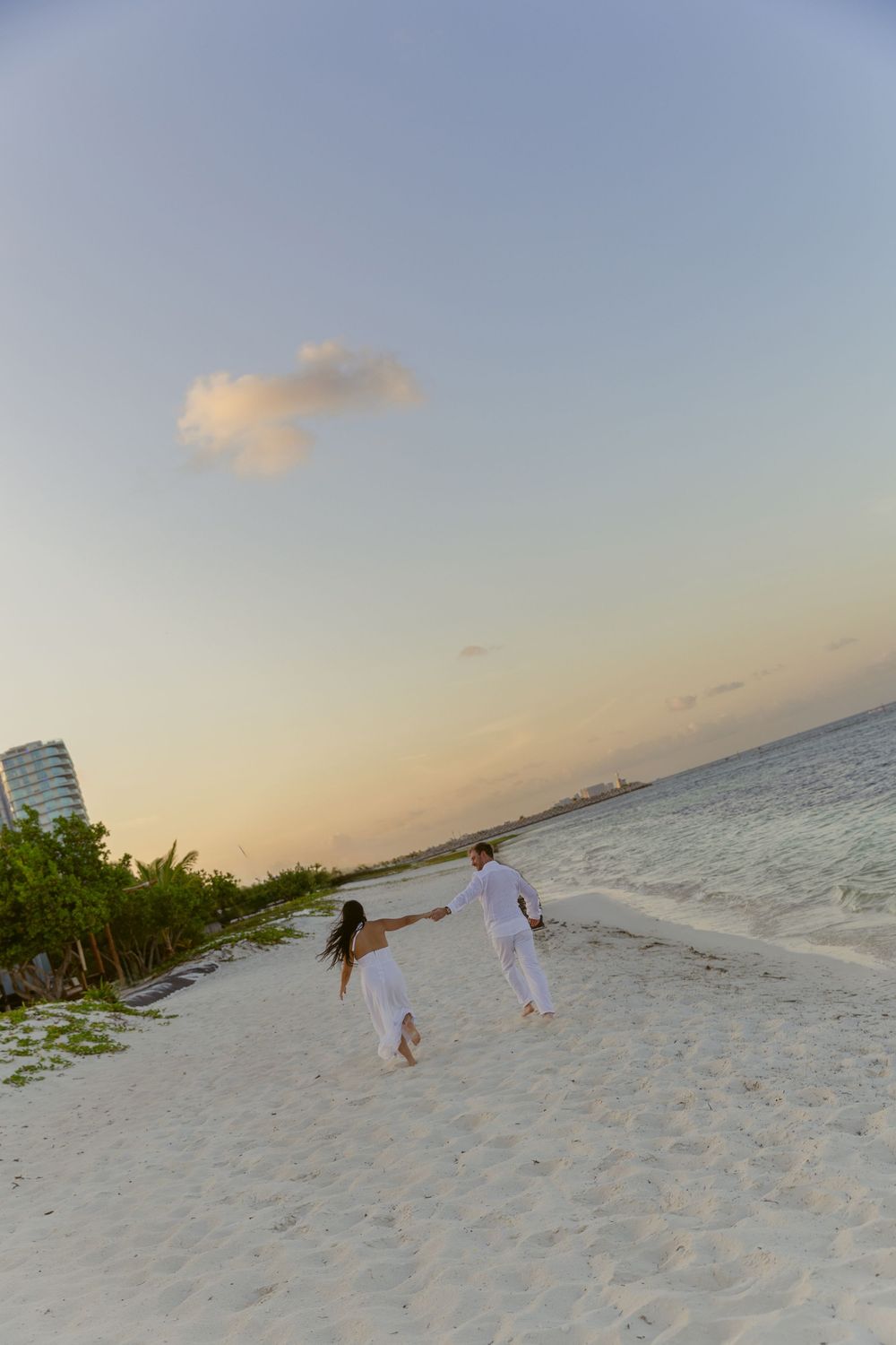Couple in white holding hands running on a white sand beach at sunset with turquoise water.