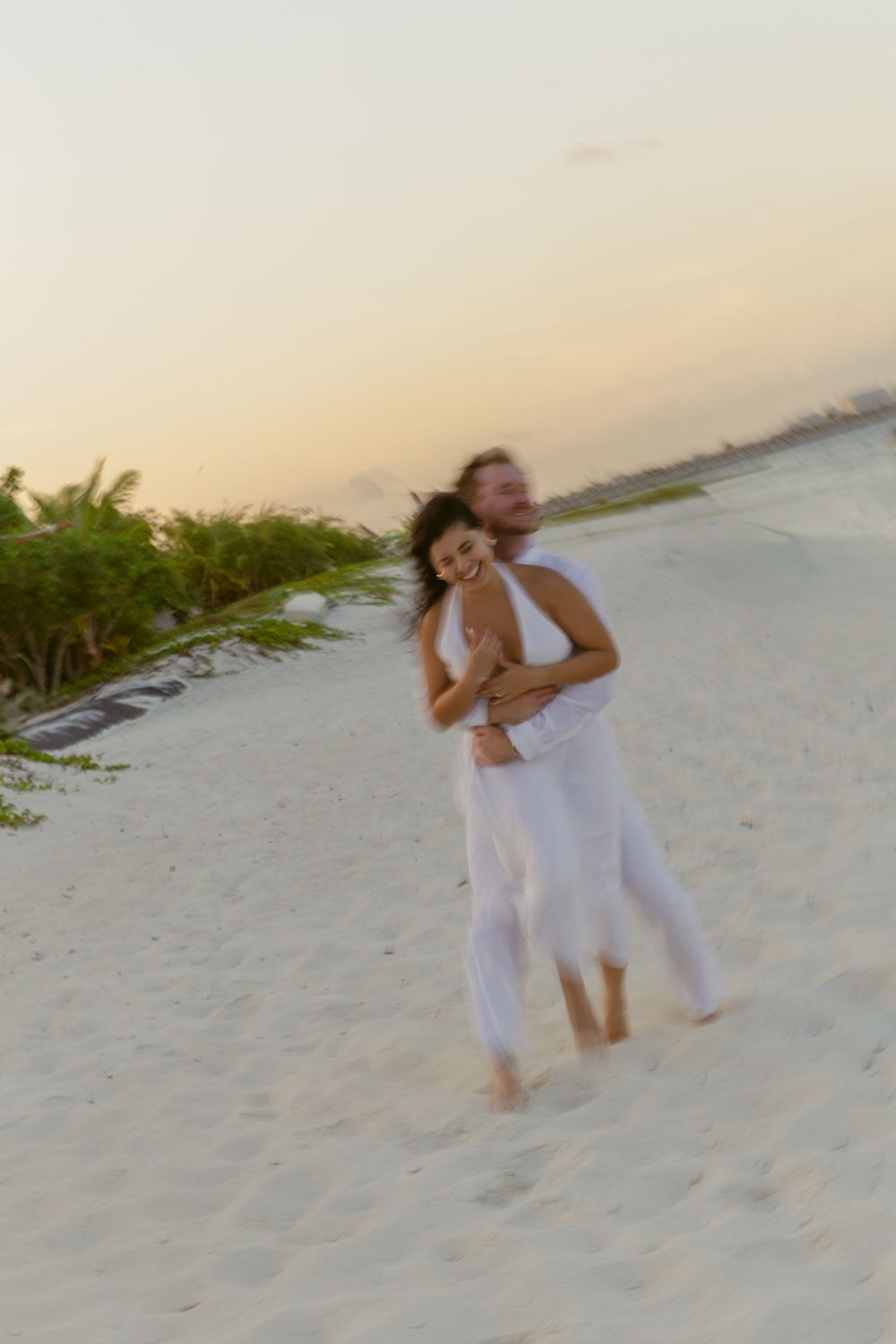 Happy couple embracing on a sandy beach at golden hour, both dressed in white outfits.