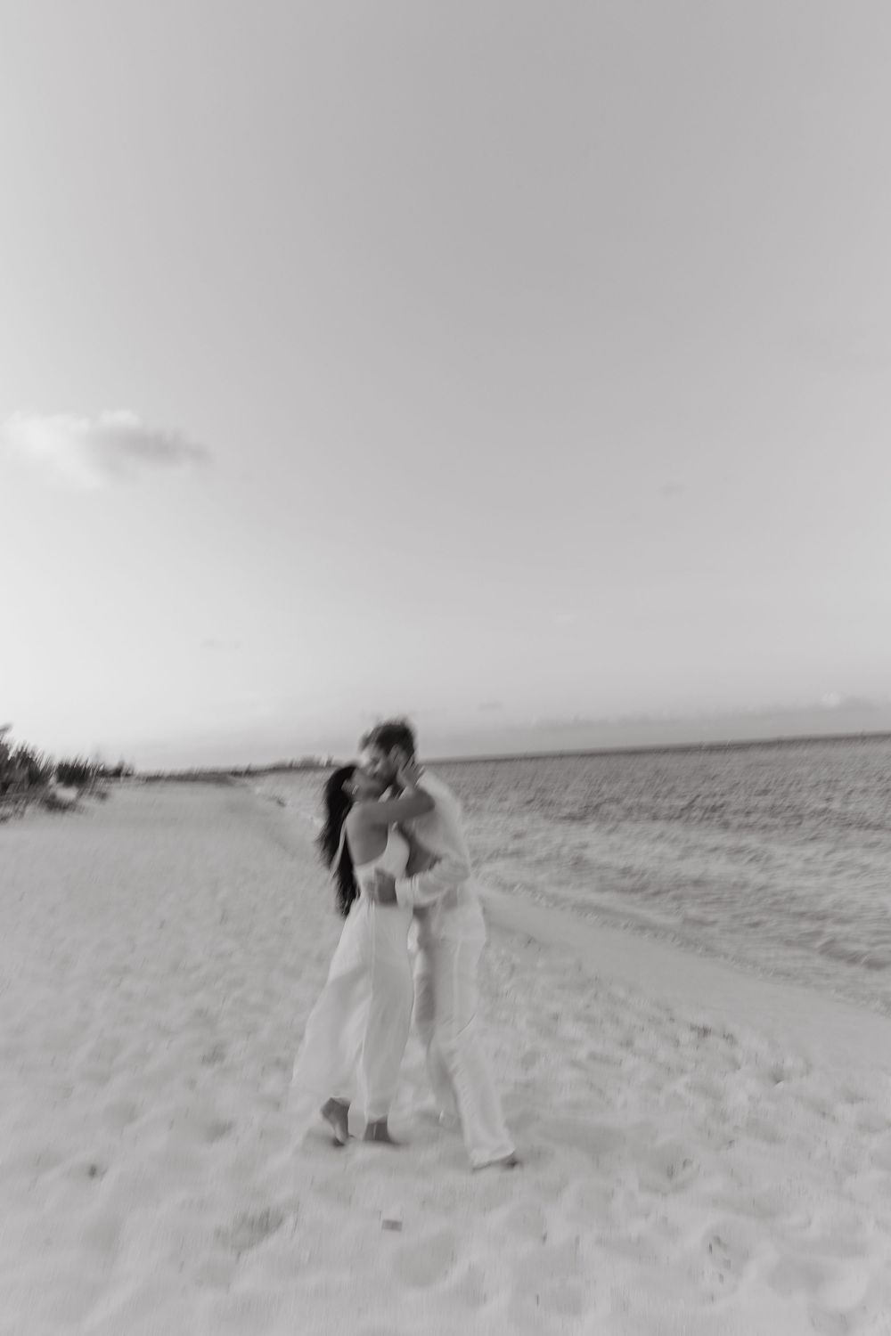Black and white photo of couple kissing on a wide sandy beach with open sky behind them.