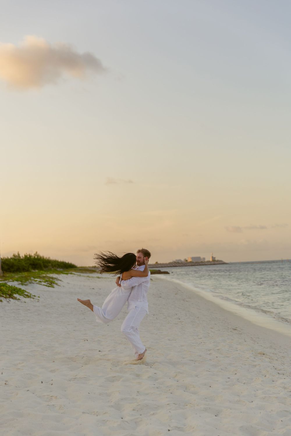 Man lifting woman in white dress on a tropical beach at sunset, her hair flowing in the wind.
