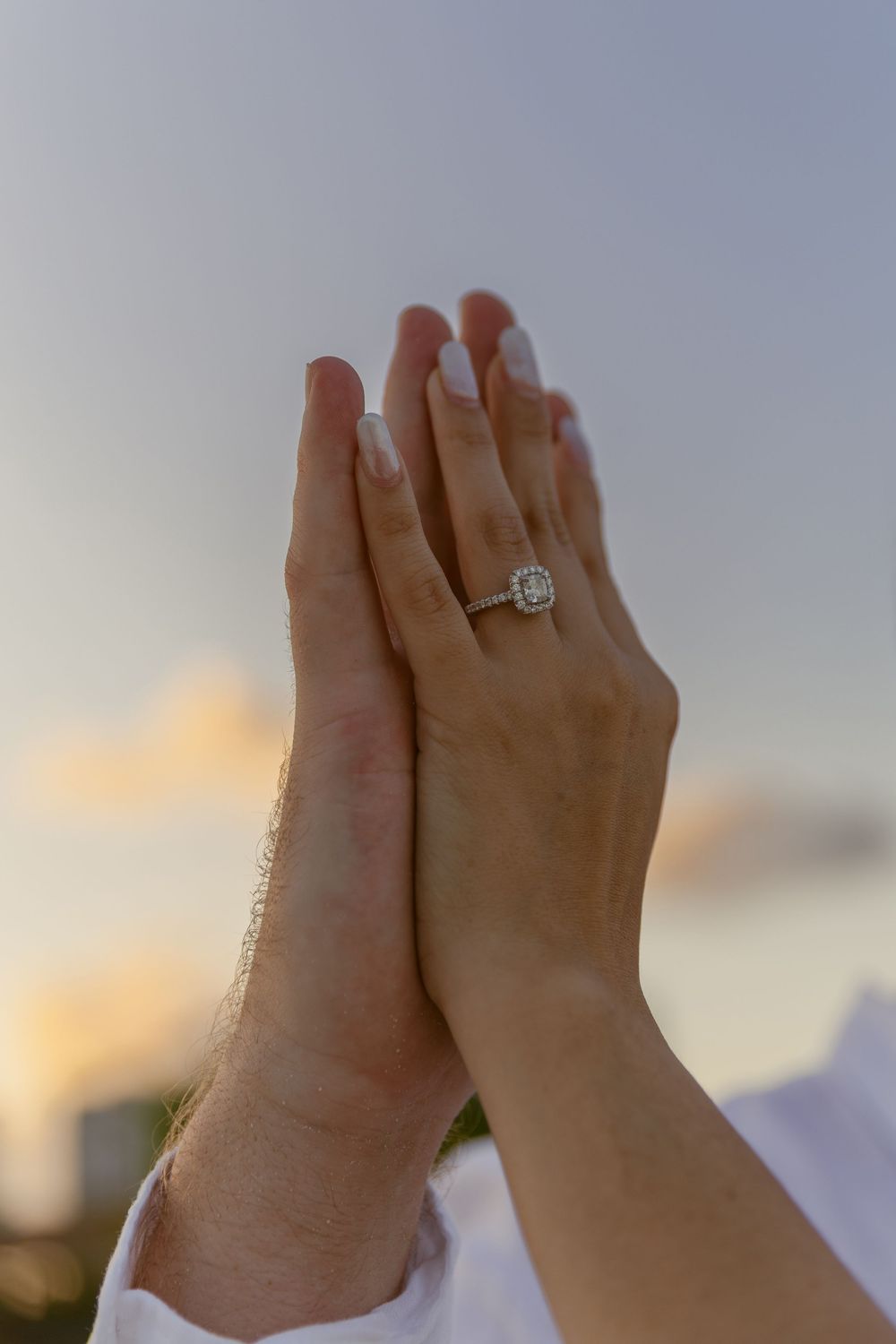 Close-up of hands pressed together displaying a diamond halo engagement ring at sunset.