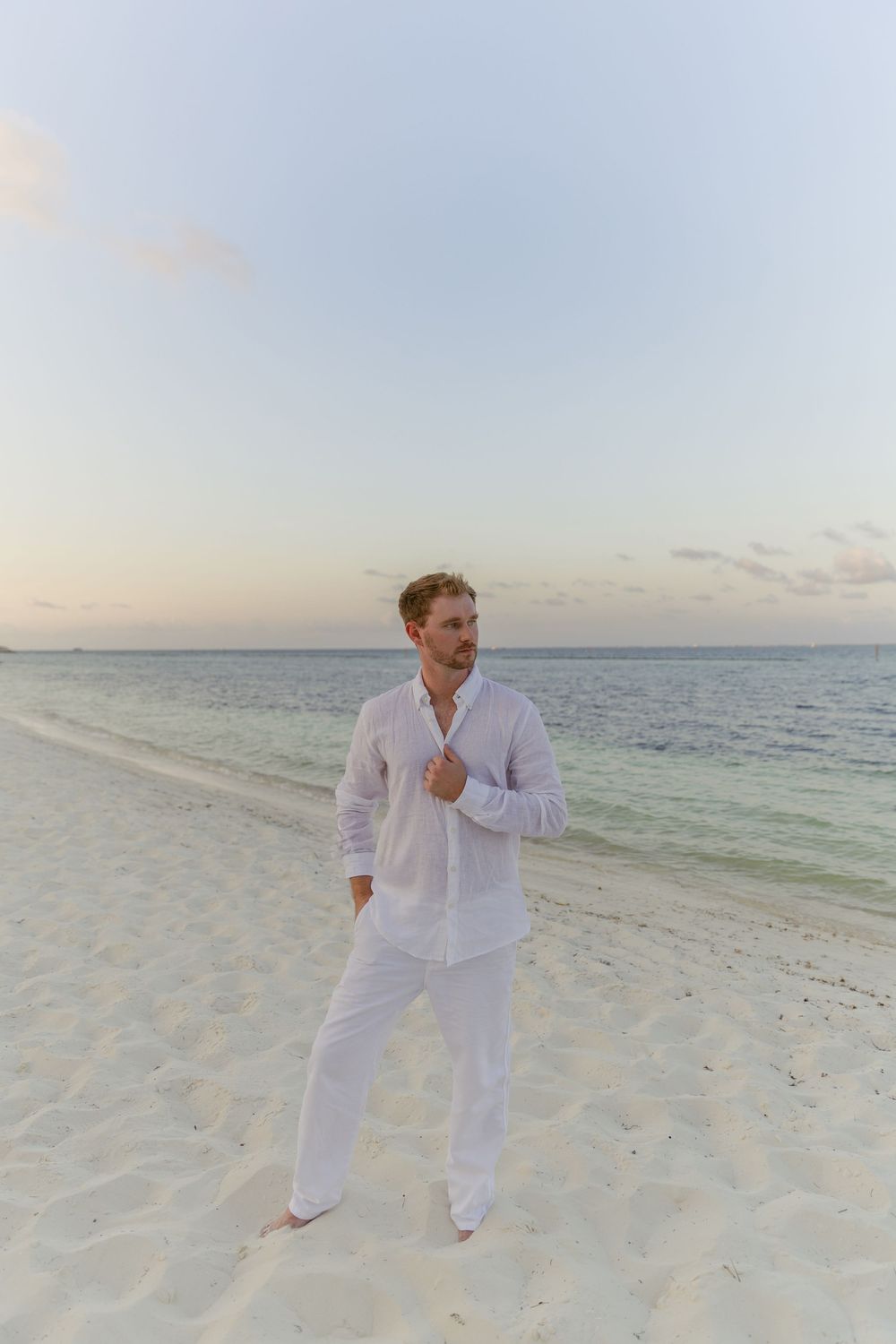 Handsome man in white linen shirt and pants standing barefoot on a beach at dusk.