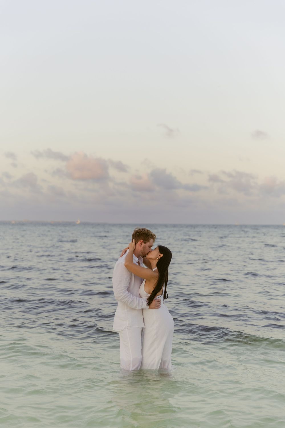 Romantic couple kissing while standing in shallow ocean water at a tropical beach at sunset.