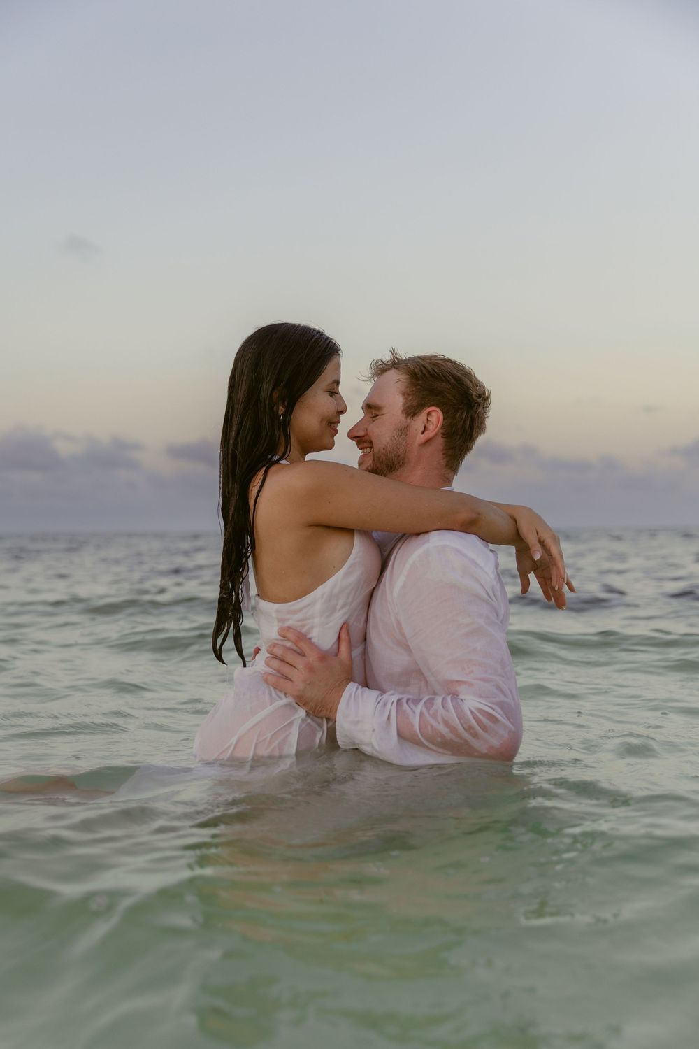 Couple embracing in the ocean with wet white clothing, smiling at each other at dusk.