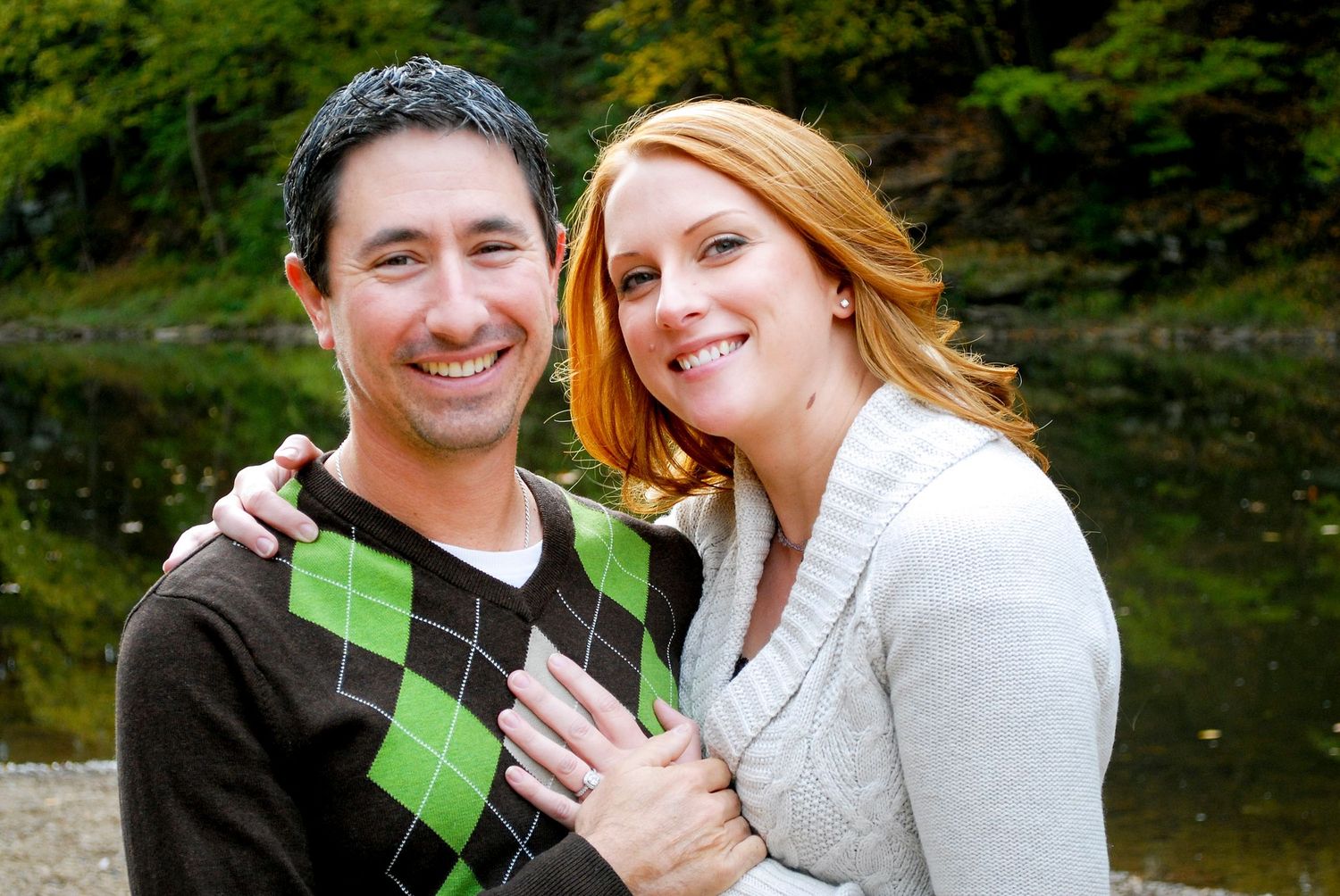 Smiling couple embracing outdoors near a pond, man in argyle sweater and woman with red hair in white cardigan.