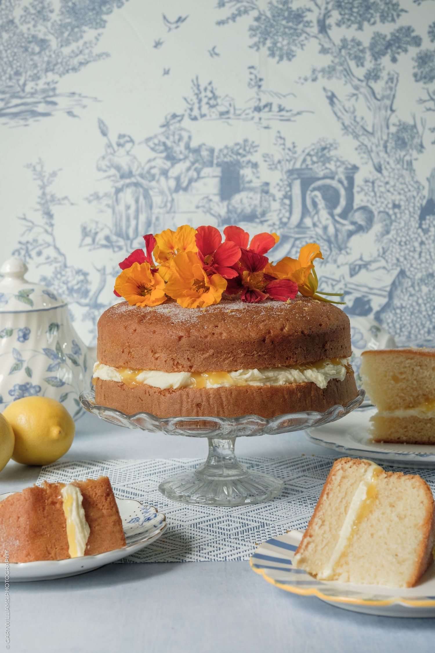 Elegant lemon Victoria sponge cake on crystal stand, topped with fresh flowers, displayed with lemons on blue toile background.