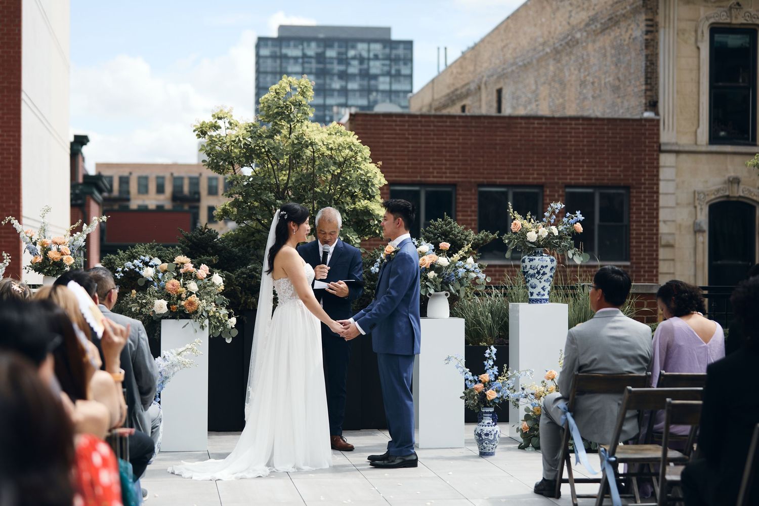 Couple exchanges vows at rooftop wedding ceremony with floral arrangements on white pedestals and city buildings in background.