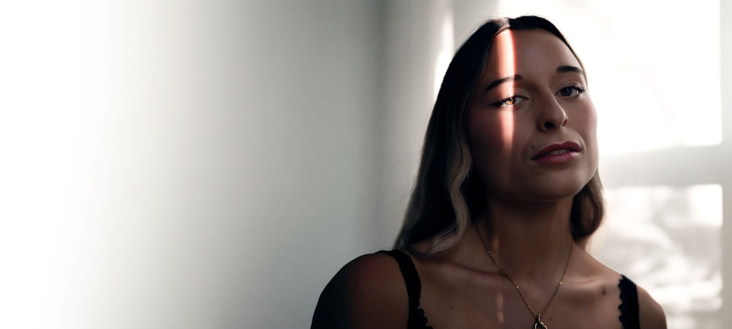 A young woman with long dark hair wearing a black top and necklace, gazing upward in soft natural light.