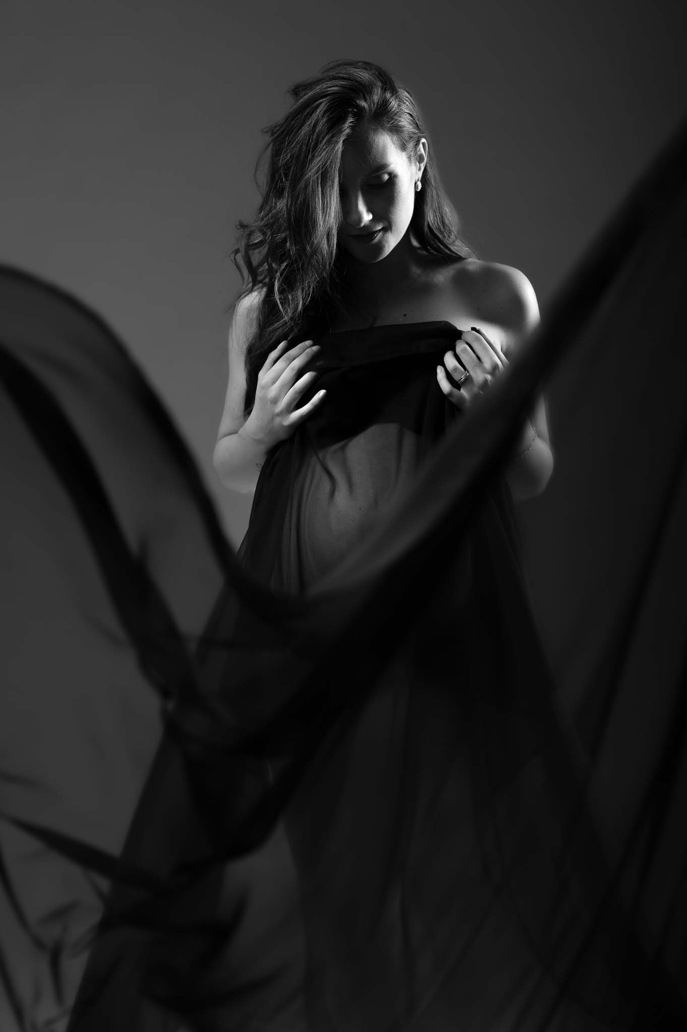 Black and white fine art portrait of a woman draped in flowing fabric, studio photography with dramatic lighting.