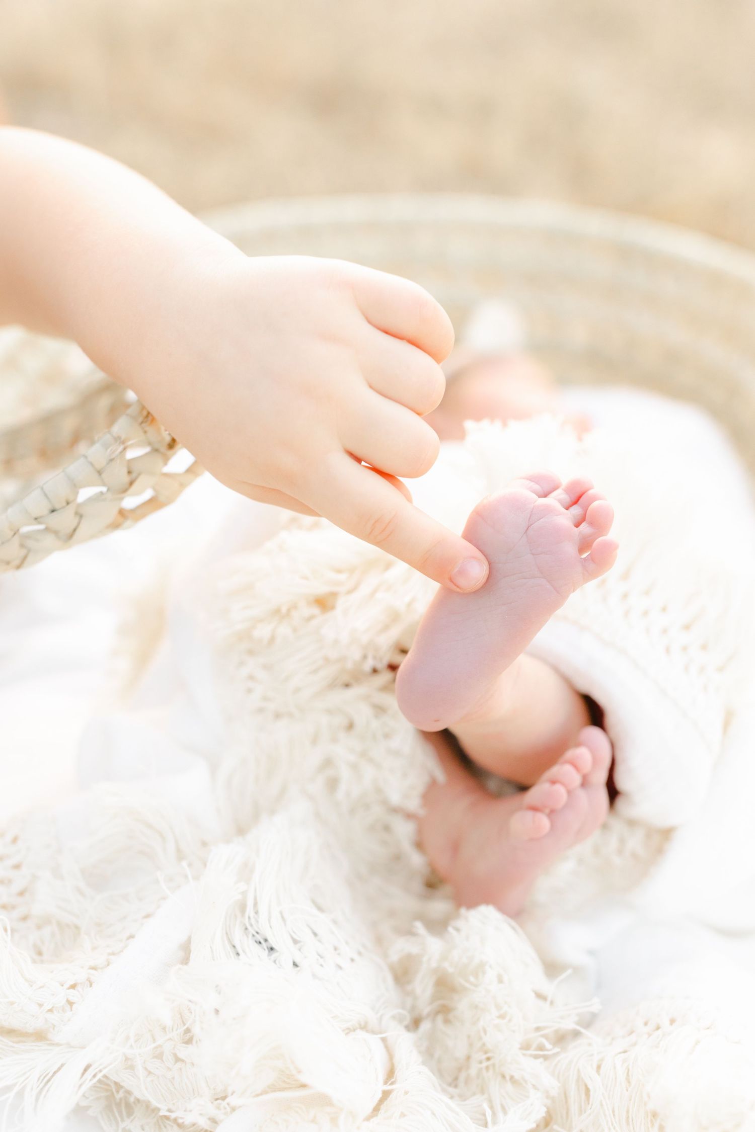 A toddler's hand gently touches tiny newborn baby feet wrapped in a soft white knit blanket in a wicker basket.