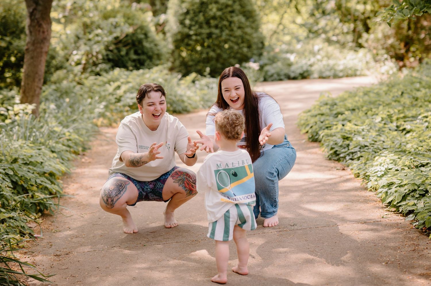 Two parents crouch on a garden path, smiling as their toddler runs toward them outdoors.