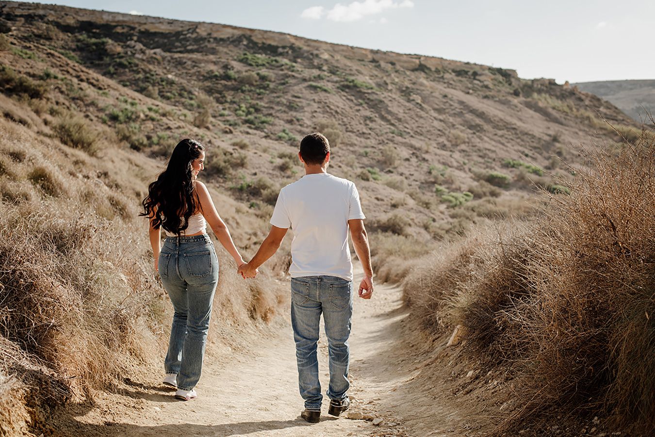 Couple holding hands walking on a dirt trail through dry golden hills in a sunny outdoor setting.