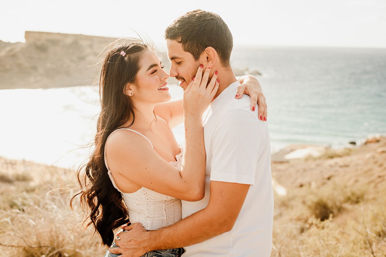 Romantic couple embracing and smiling near a scenic coastal cliff with ocean views on a sunny day.
