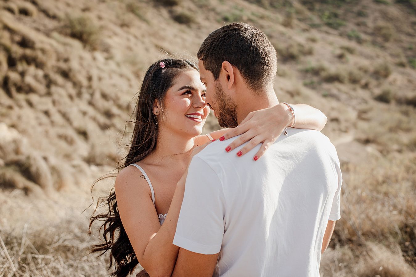 A smiling woman with long dark hair embraces a man outdoors in a sunny, arid landscape.