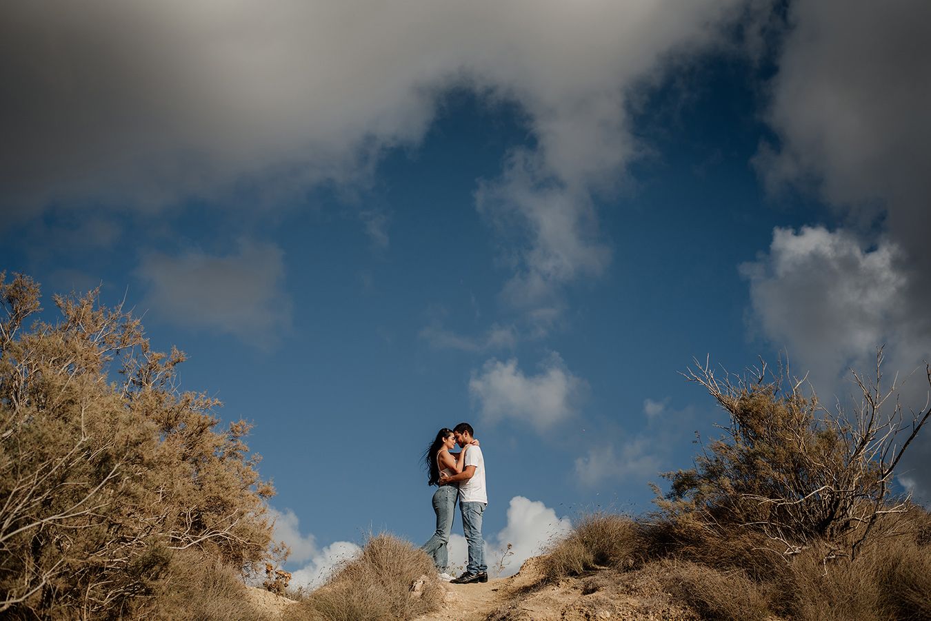 Couple embracing on a rocky hilltop surrounded by desert shrubs under a dramatic blue sky with clouds.
