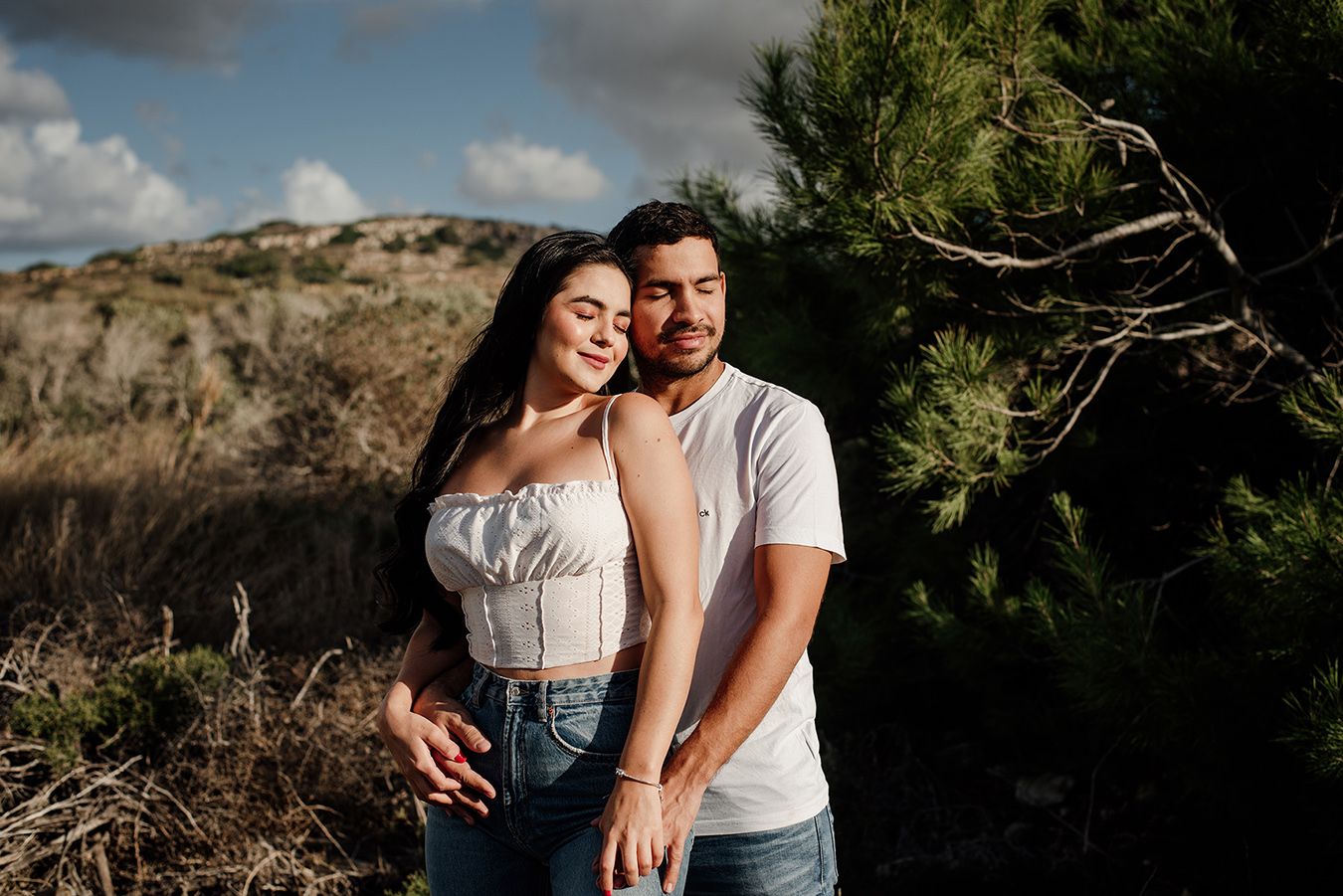 Couple embracing outdoors in nature, woman in white crop top and man in white tee, with green trees and hills behind.