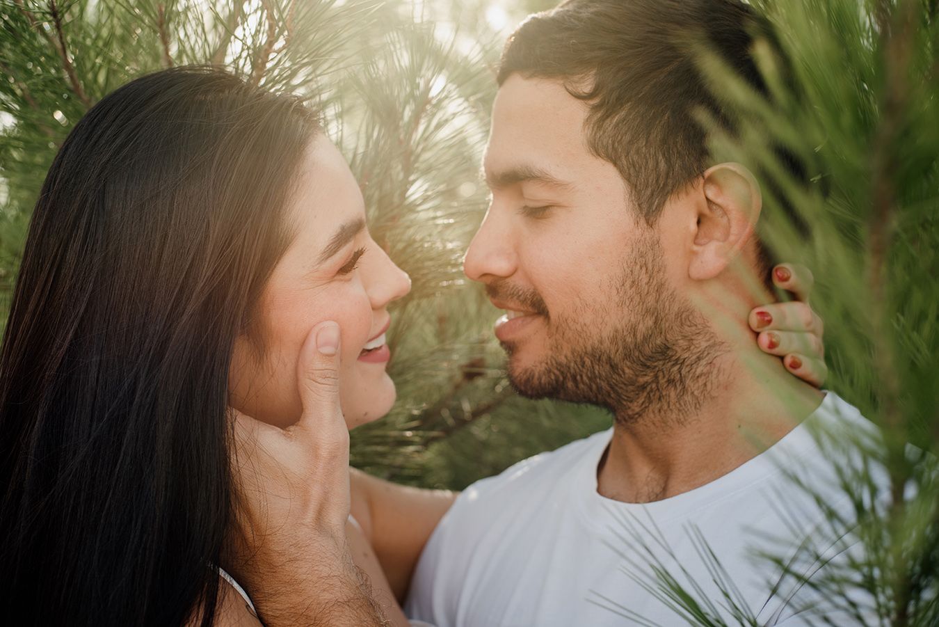 A romantic couple sharing a tender moment outdoors, surrounded by lush green pine trees in soft natural light.