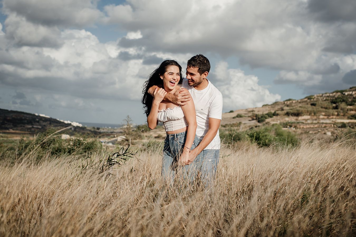 Happy couple embracing in a golden field with cloudy sky and hills in the background.