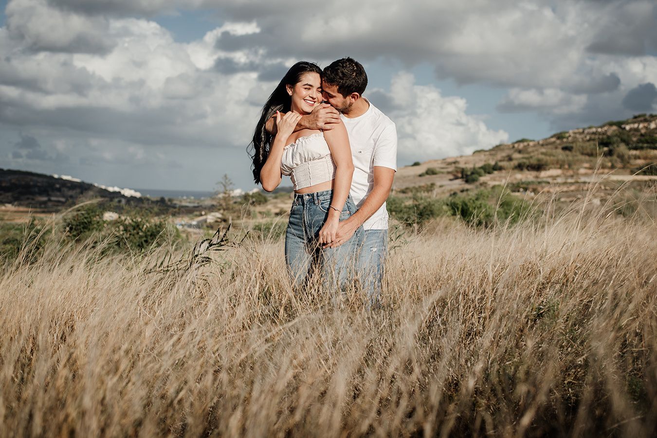 Couple embracing romantically in a golden wheat field under dramatic cloudy skies in a scenic outdoor engagement photo.