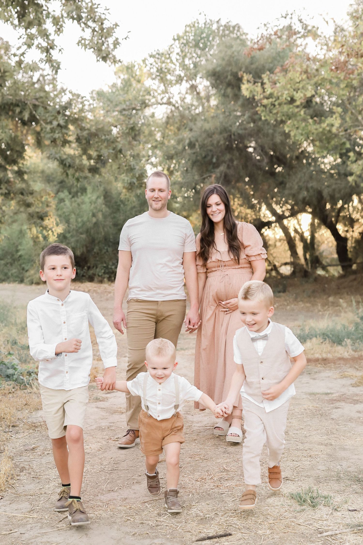 Pregnant woman in pink dress with husband and three young boys walking outdoors during a family maternity photo session.