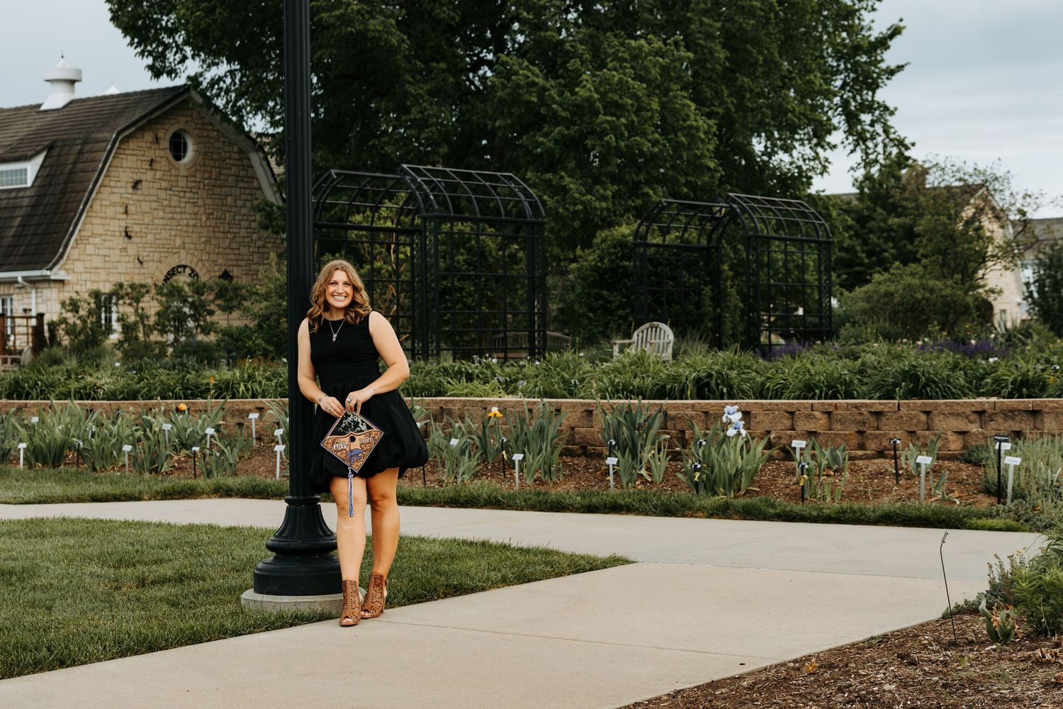 Woman in black dress posing by a lamppost in a lush garden with a stone building backdrop.