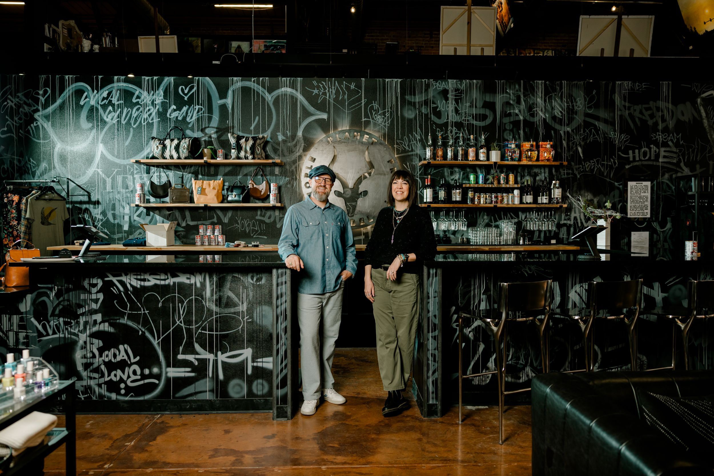 Two people standing behind a bar with graffiti walls and shelves of bottles in a trendy urban cocktail bar.