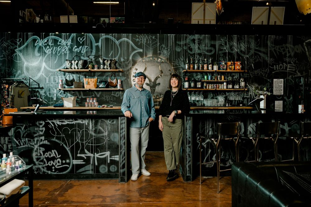 Two people standing behind a bar with graffiti walls and shelves of bottles in a trendy urban cocktail bar.
