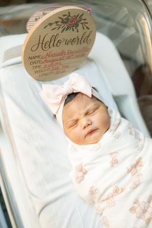 Newborn baby girl swaddled in floral blanket with pink bow headband, sleeping peacefully in hospital bassinet with Hello World sign.