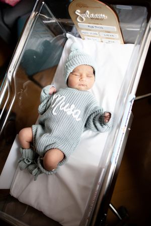 Newborn baby sleeping in hospital bassinet wearing a mint green knit outfit and matching hat with name embroidered at Fort Sanders.