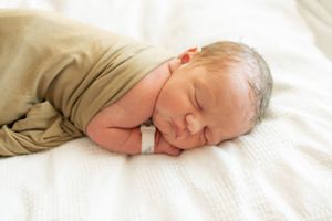 Peaceful newborn baby swaddled in olive green wrap sleeping on white blanket with hospital bracelet.