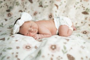 Newborn baby sleeping peacefully on floral blanket wearing white bow headband and diaper.