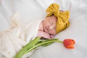 Sleeping newborn baby wrapped in white swaddle wearing yellow bow headband beside a red tulip.