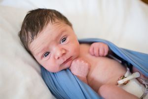 Newborn baby wrapped in a soft blue swaddle blanket, with dark hair and alert eyes, resting on white bedding.