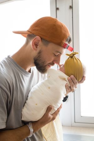 A bearded man in an orange cap tenderly kisses his newborn baby wrapped in white swaddle cloth near a bright window.
