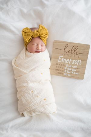 Newborn baby girl named Emerson swaddled in white muslin wrap with yellow bow headband beside birth announcement sign.