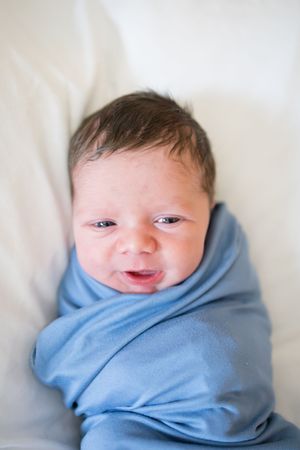 Newborn baby swaddled in blue blanket with dark hair, making an alert expression while lying on white background.