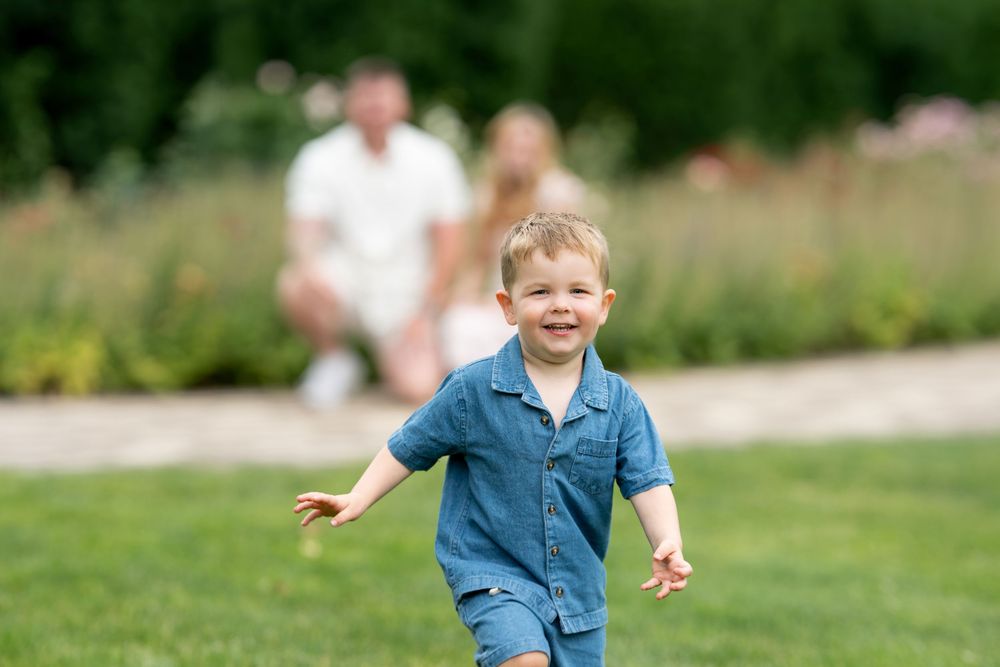 A happy toddler boy in denim outfit runs on green grass while his parents watch in the background.
