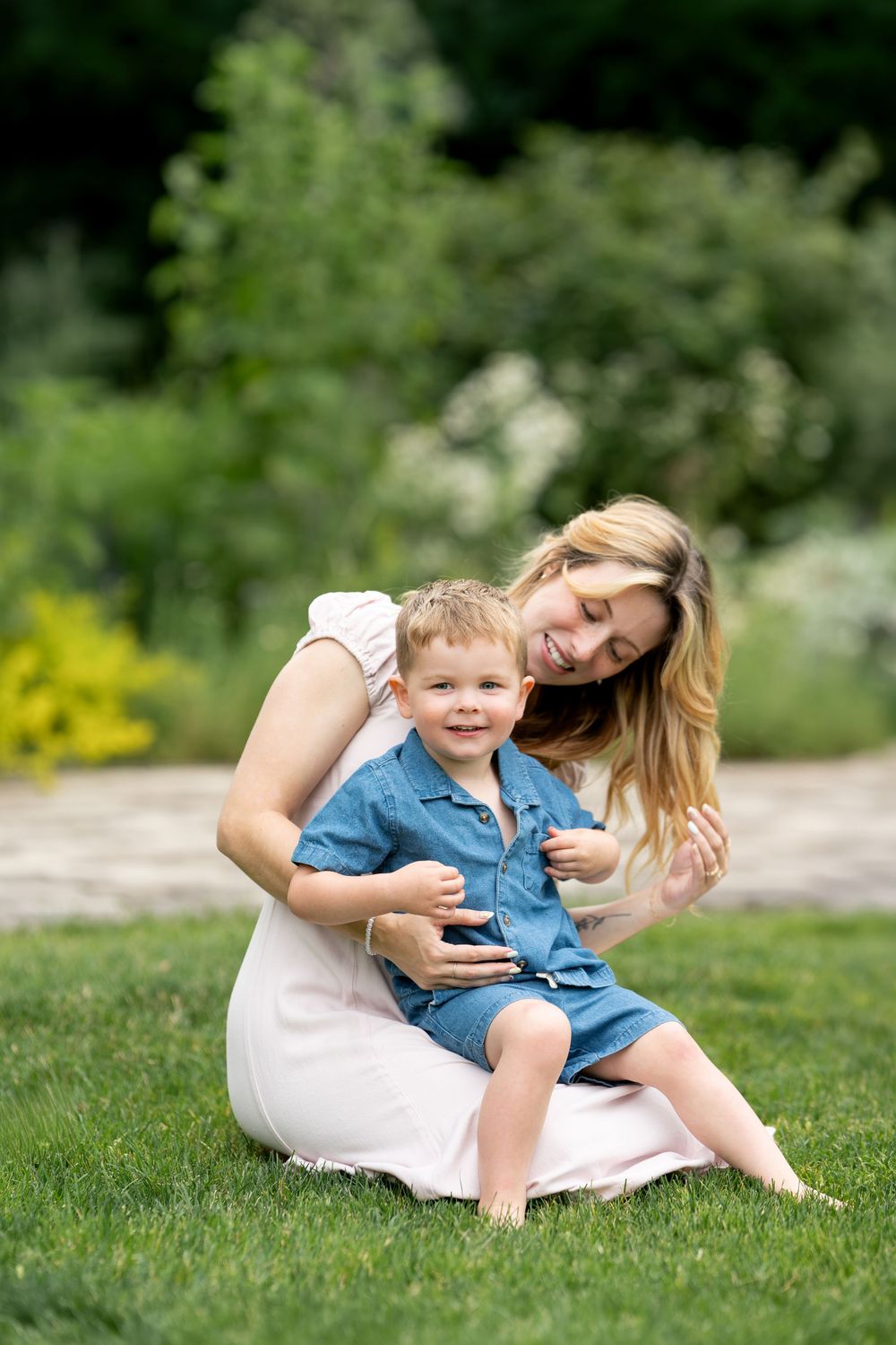 Smiling mother sitting barefoot on green grass holding her happy toddler son in a sunny garden.