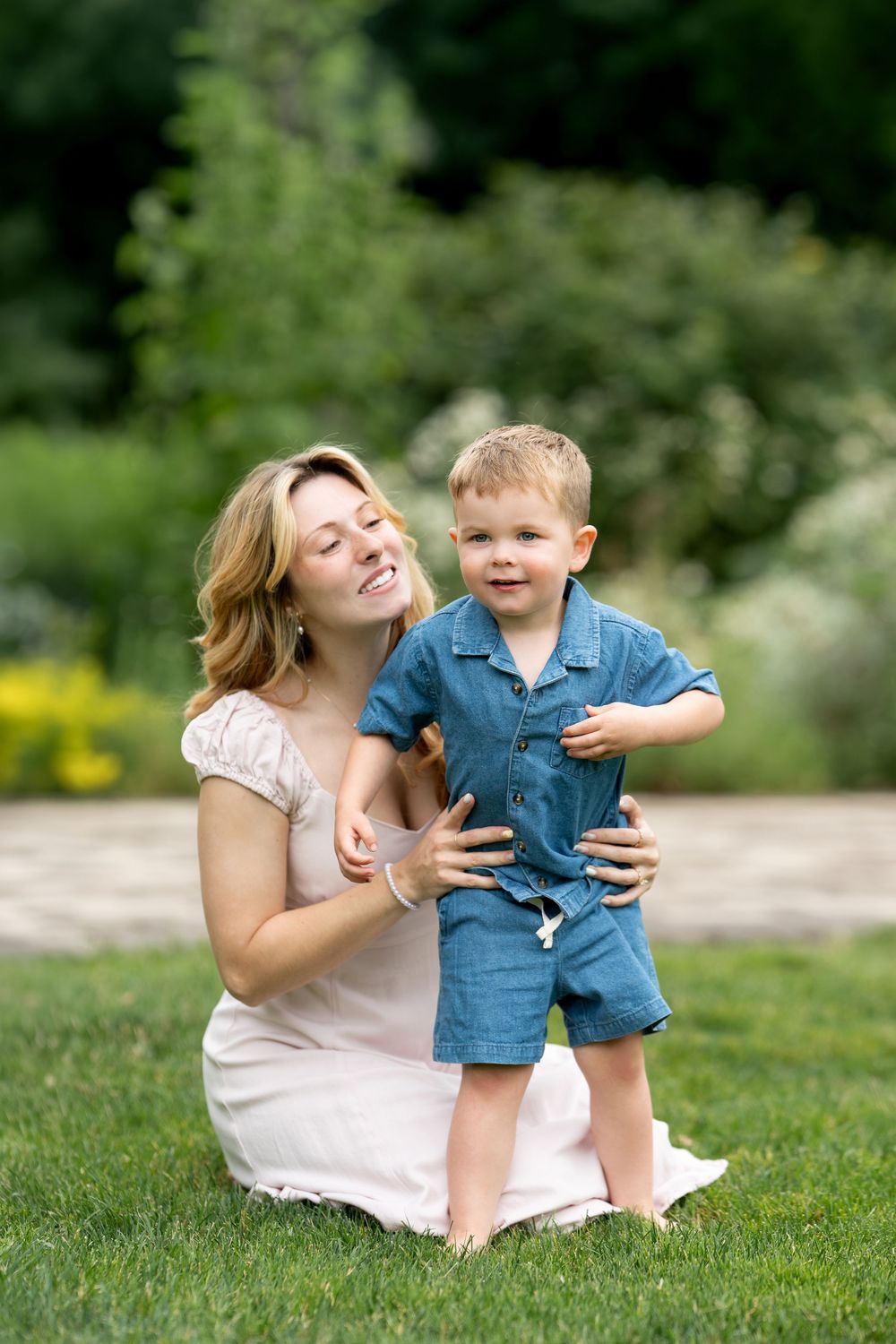 A smiling mother in a pink dress sits on grass holding her toddler son in a blue denim outfit outdoors.