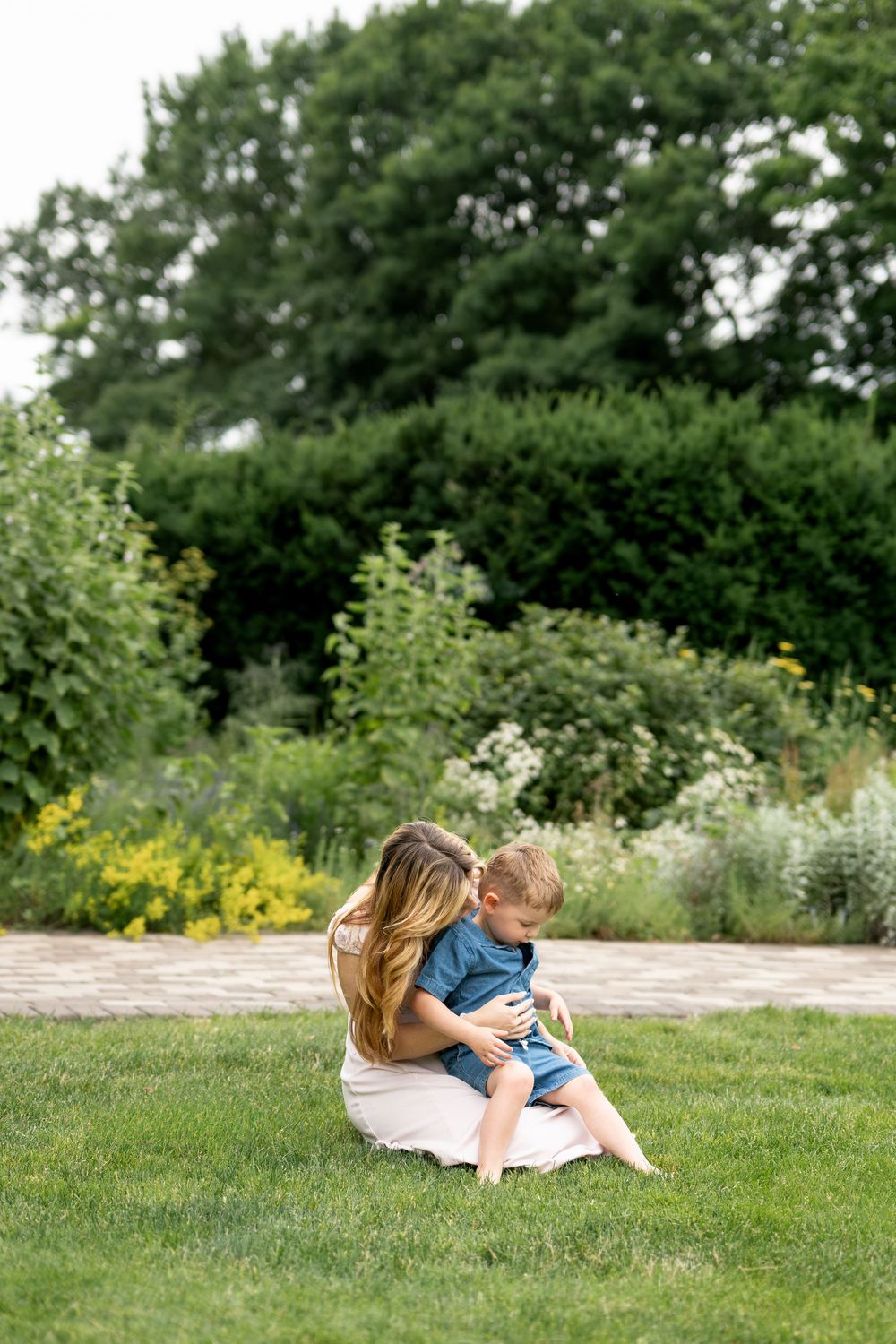 A loving mother sits on grass hugging her young son in a lush garden setting.