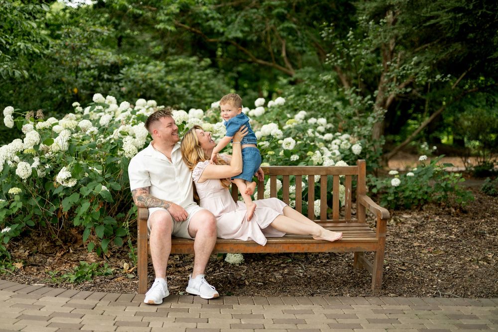Happy family of three sitting on a wooden bench surrounded by white hydrangeas in a garden.