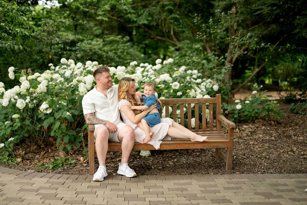 A happy family of three sits on a wooden bench surrounded by white hydrangeas in a beautiful garden.