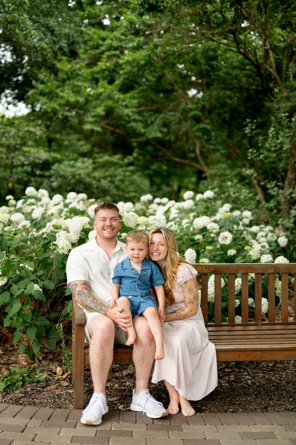 Family of three sitting on wooden bench surrounded by white hydrangeas in a green garden.