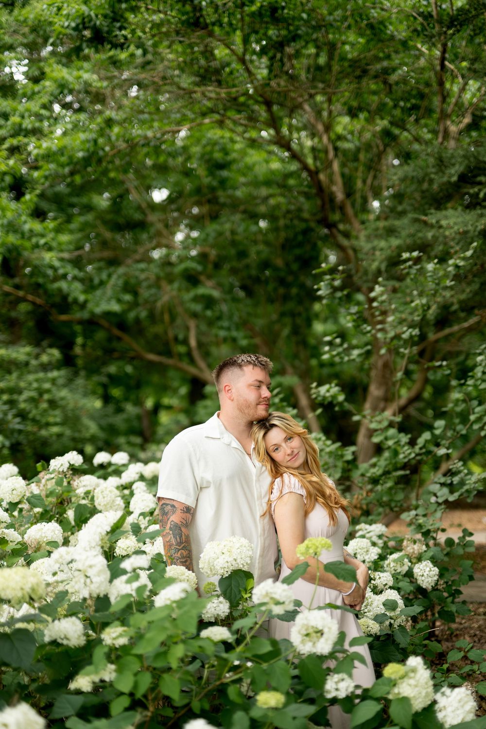 A couple stands together surrounded by white hydrangeas and green trees in a serene garden.